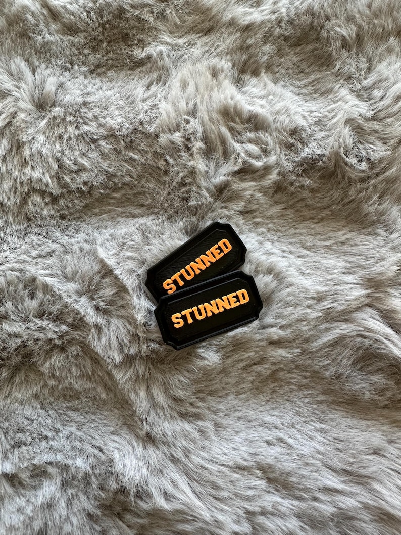 Puede incluir: Dos parches rectangulares negros con la palabra "STUNNED" en letras naranjas. Los parches est&aacute;n sobre una superficie gris y esponjosa.