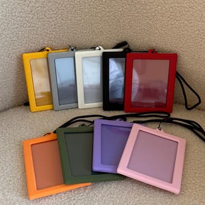Könnte beinhalten: Eine Sammlung farbenfroher Ausweishalter in verschiedenen Farbtönen, darunter Gelb, Grau, Weiß, Schwarz, Rot, Orange, Grün, Lila und Rosa. Jeder Halter hat ein klares Fenster und ein schwarzes Umhängeband.