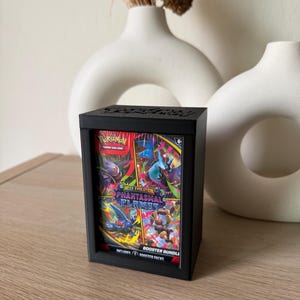 May include: A black display box holding a Pokemon trading card game booster bundle. The bundle features the "Phantasmal Flames" set with colorful graphics of Pokemon characters. The box is on a wooden surface.