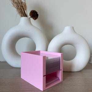 Op de afbeelding: Een roze, vierkante bureau-organizer met twee compartimenten, met stapels witte kaarten. Twee witte, cirkelvormige vazen met een gat in het midden staan op de achtergrond. Gedroogde bruine planten staan in een vaas.