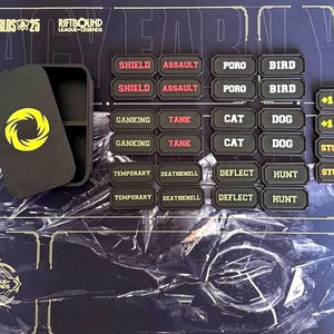 May include: A black game piece set with various text labels, including "SHIELD," "ASSAULT," and "GANKING." A black box with a yellow spiral logo is open, revealing compartments. The game board has the text "WORLDS 25" and "RIFTBOUND LEAGUE-LEGENDS."