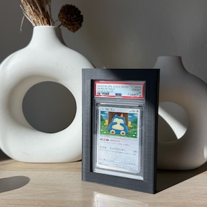 Może przedstawiać: Oprawiona karta kolekcjonerska Pokémon z Snorlax z zestawu Sun & Moon Double Blaze. Karta oceniona na GEM MT 10 przez PA. Czarna ramka prezentuje kartę na drewnianej powierzchni, z białymi wazonami w tle.