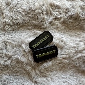 Puede incluir: Dos parches rectangulares negros con la palabra "TEMPORARY" en letras verdes. Los parches tienen un borde decorativo y est&aacute;n colocados sobre un fondo gris y esponjoso.