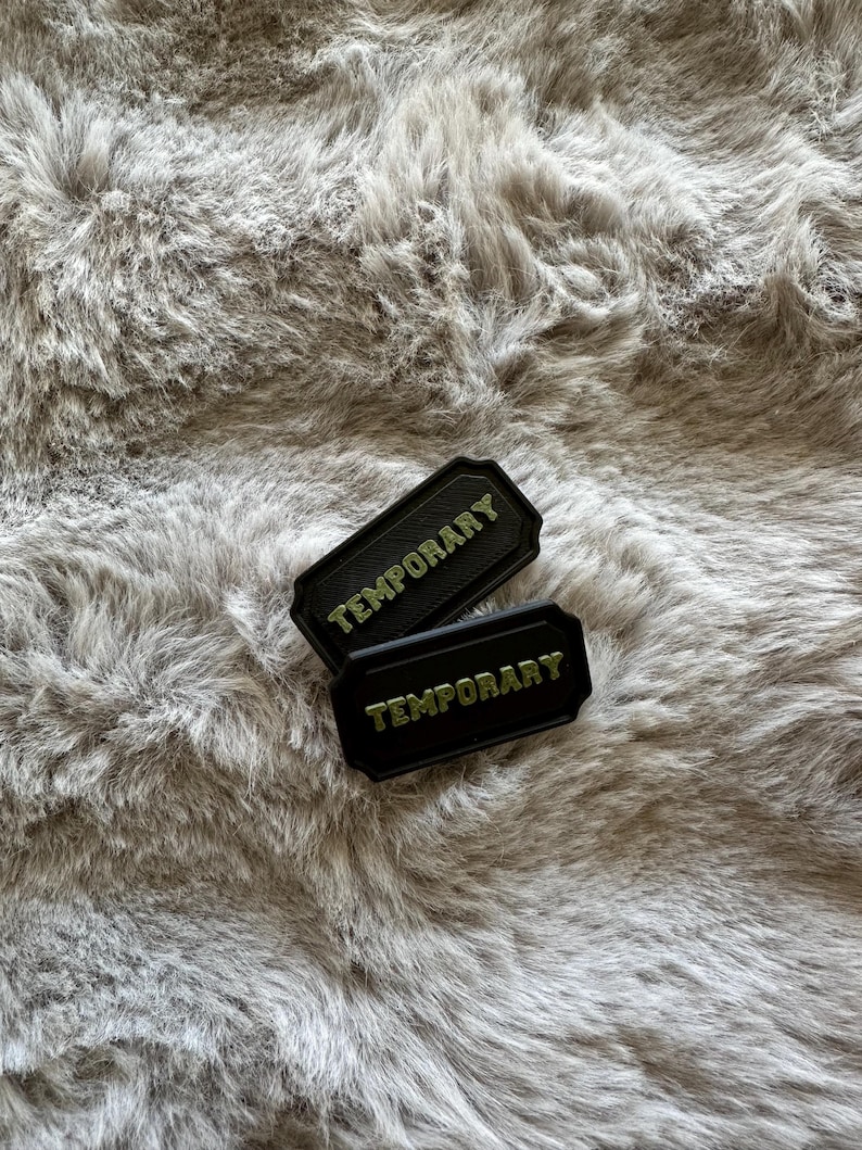 Puede incluir: Dos parches rectangulares negros con la palabra "TEMPORARY" en letras verdes. Los parches tienen un borde decorativo y est&aacute;n colocados sobre un fondo gris y esponjoso.
