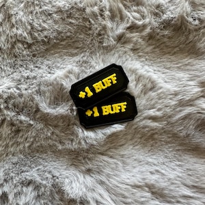 Puede incluir: Dos fichas rectangulares negras con texto amarillo "+1 BUFF" se muestran sobre una superficie gris y esponjosa. Las fichas tienen un borde decorativo. El fondo es un material suave y texturizado.