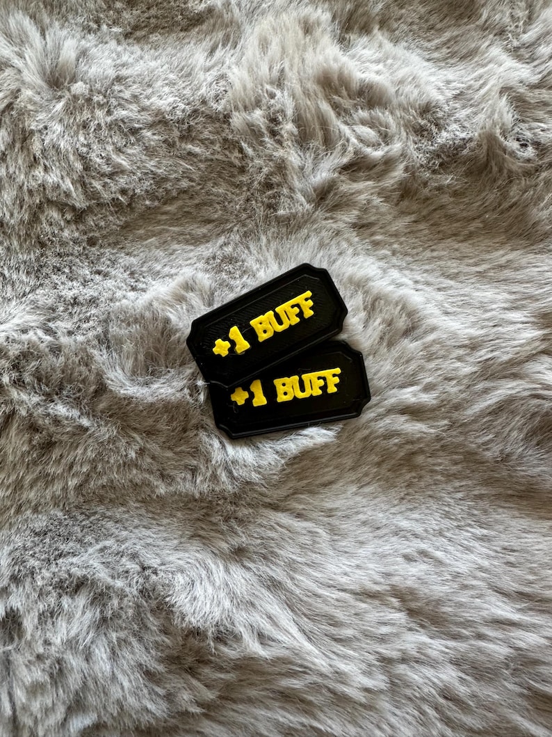 Puede incluir: Dos fichas rectangulares negras con texto amarillo "+1 BUFF" se muestran sobre una superficie gris y esponjosa. Las fichas tienen un borde decorativo. El fondo es un material suave y texturizado.