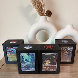 May include: Four black display boxes with Pokémon trading cards. Each box has a different Pokémon card visible through a clear window. The boxes are decorated with Pokémon-themed logos on top.