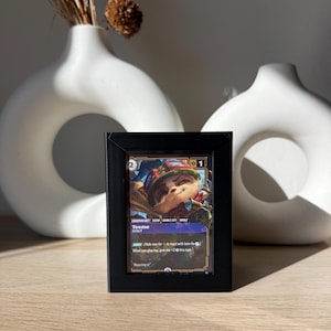 May include: A framed card featuring the character Teemo from the game League of Legends. The card is in a black frame, with the character's name and abilities visible. The card is displayed on a wooden surface, with white vases in the background.