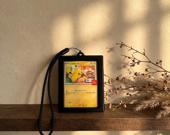 May include: A black plastic card holder containing a yellow Pikachu trading card with Japanese text and a small red McDonald's logo. The card holder features a black lanyard attached to the top, allowing for easy carrying or display of the collectible.