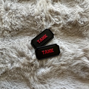 Puede incluir: Dos parches rectangulares negros con la palabra "TANK" en letras rojas. Los parches tienen un borde en relieve y est&aacute;n colocados sobre un fondo gris y esponjoso.