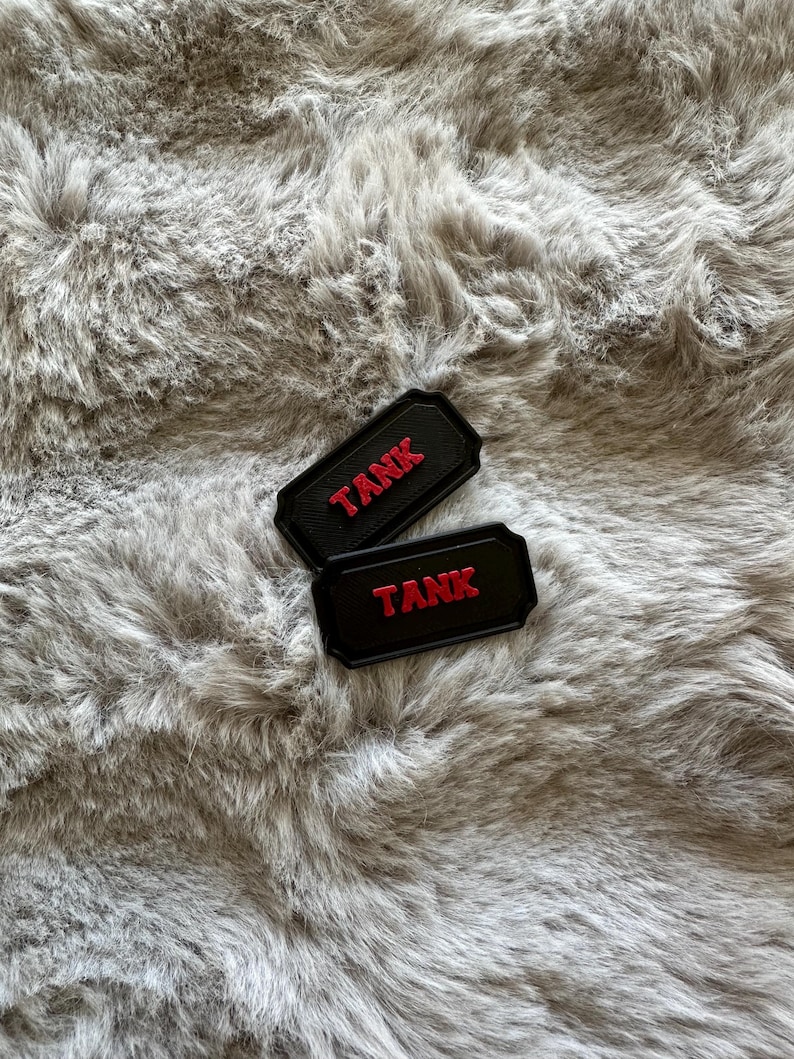 Puede incluir: Dos parches rectangulares negros con la palabra "TANK" en letras rojas. Los parches tienen un borde en relieve y est&aacute;n colocados sobre un fondo gris y esponjoso.