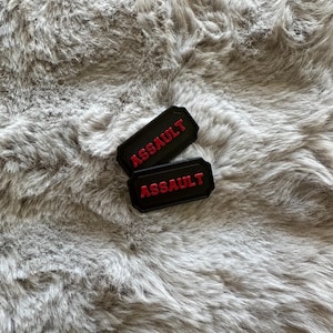 Puede incluir: Dos parches rectangulares negros con la palabra "ASSAULT" en letras rojas. Los parches se muestran sobre una superficie gris texturizada.