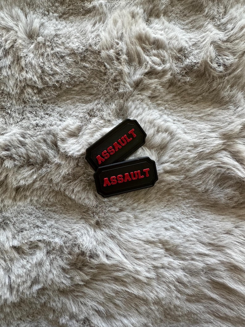 Puede incluir: Dos parches rectangulares negros con la palabra "ASSAULT" en letras rojas. Los parches se muestran sobre una superficie gris texturizada.