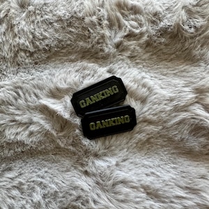 Puede incluir: Dos parches rectangulares negros con la palabra "GANKING" en letras doradas. Los parches est&aacute;n colocados sobre un fondo de material de piel sint&eacute;tica gris suave. Los parches tienen un borde texturizado.