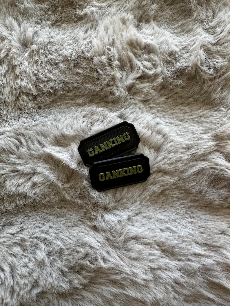 Puede incluir: Dos parches rectangulares negros con la palabra "GANKING" en letras doradas. Los parches est&aacute;n colocados sobre un fondo de material de piel sint&eacute;tica gris suave. Los parches tienen un borde texturizado.