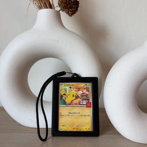 Op de afbeelding: Een zwarte kaarthouder met een zwart koord bevat een Pokémon-kaart met Pikachu. De kaart heeft Japanse tekst en een McDonald's-logo. De achtergrond omvat witte keramische vazen en gedroogde bloemstukken.