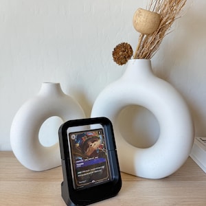 May include: A black card display case holding a trading card with the name "Teemo" visible. The case is on a wooden surface, with two white, ring-shaped vases and dried floral arrangements in the background.