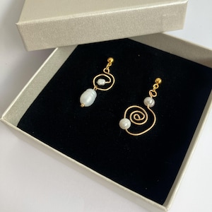 Può includere: Un paio di orecchini in filo color oro con perle bianche in un design a spirale, presentati in una scatola beige con l'etichetta GRETA ART & JEWELRY. Gli orecchini hanno un perno a lobo e un elemento a perla pendente con finitura metallica.