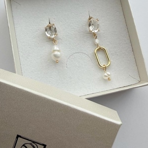 Può includere: Un paio di orecchini spaiati con pietre ovali trasparenti, piccole perle bianche e un anello ovale color oro. Gli orecchini sono presentati in una scatola bianca con il testo GRETA ART & JEWELRY stampato in un logo quadrato nero sul coperchio.