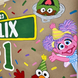 Peut inclure: Panneau en papier brun avec les personnages de Sesame Street, Elmo, Abby Cadabby et Oscar le Grognon. Le panneau porte le nom "Felix" et le chiffre "1" en blanc et vert. Des confettis color&eacute;s et un bord jaune compl&egrave;tent le th&egrave;me de l'anniversaire.