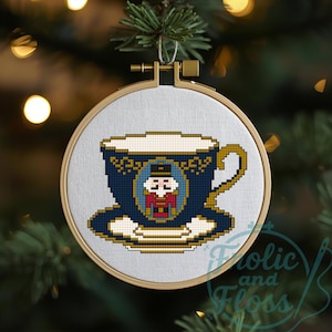 May include: A cross-stitch Christmas ornament featuring a blue teacup with a gold handle and trim, and a nutcracker design. The ornament is displayed in a wooden embroidery hoop, hanging from a Christmas tree.
