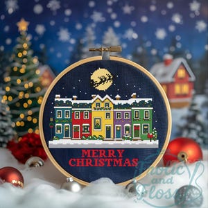 May include: A cross-stitch hoop art piece depicting a row of colourful houses with snow-covered roofs. The design includes a full moon with Santa's sleigh and the text "MERRY CHRISTMAS" in red. The hoop is set against a snowy background.