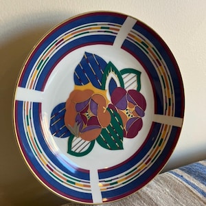 Könnte beinhalten: Ein dekorativer Teller mit floralem Design. Der Teller hat eine weiße Mitte mit orangefarbenen und lila Blumen und grünen Blättern. Der Rand hat ein blaues, rotes, gelbes und grünes Streifenmuster mit goldenem Rand. Weiße Linien kreuzen den Rand.