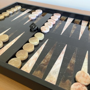 Puede incluir: Un tablero de backgammon con una superficie de juego negra y puntas triangulares incrustadas en tonos marrón y crema. Las fichas son de color marrón claro y rosa, y un dado negro muestra el número 16.