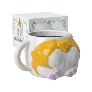 以下が含まれることがあります： 白いセラミックマグカップ。コーギーのお尻のデザインで、黄色と白の毛並みとピンクの足が特徴です。マグカップには濃い液体が入っており、カーブしたハンドルが付いています。マグカップは、「CORGI PALACE」の文字が入った白い箱の前にあります。