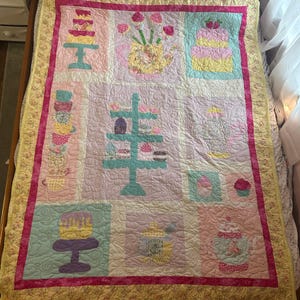 Puede incluir: Un edredón hecho a mano con un diseño de patchwork que presenta pasteles, tazas de té y teteras en colores pastel. El edredón tiene un borde rosa y un borde con estampado floral.
