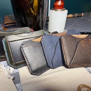 May include: A collection of handmade leather cardholders in various colors, including green, black, and brown. Each wallet features contrasting stitching and a unique design, showcasing the craftsmanship. The wallets are displayed on a work surface.