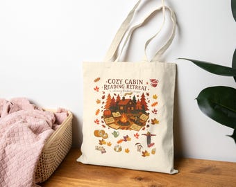 May include: Cream tote bag featuring a cabin scene with a campfire, pumpkins, and autumn leaves. The text on the bag reads "COZY CABIN READING RETREAT" and "a relaxing bookish escape". The bag has two handles and is on a wooden surface. The design is autumnal.