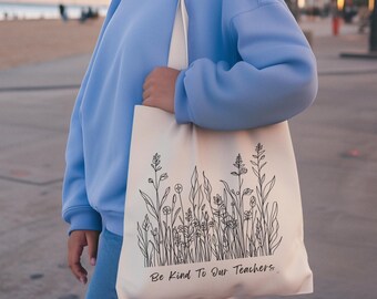 Op de afbeelding: Crèmekleurige draagtas met een zwart bloemenontwerp en de tekst "Be Kind To Our Teachers". De tas wordt gedragen door een persoon die een lichtblauwe trui en een spijkerbroek draagt. De achtergrond toont een strand en palmbomen.