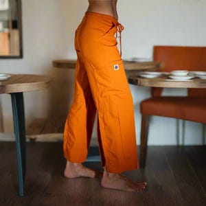 May include: Bright orange wide-leg pants with a drawstring waist and a small pocket detail. The pants are made of a lightweight fabric and have a relaxed fit. The pants are shown in a side view.