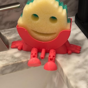 May include: A yellow sponge with a green top, held by a red plastic holder with a smiling face design. The holder has arms and legs, giving the sponge a playful appearance. The sponge is designed for cleaning.