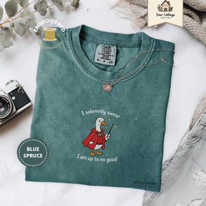 May include: Blue spruce t-shirt with an embroidered design of a duck in a red cloak holding a wand. The text "I solemnly swear I am up to no good" is above and below the duck. A silver necklace and a vintage camera are also visible.