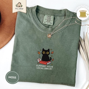 Puede incluir: Camiseta bordada verde musgo con un gato negro sentado en un libro con el texto "Support Your Local Library". El diseño incluye una taza de café y hojas de otoño. La camiseta tiene cuello redondo y mangas cortas.