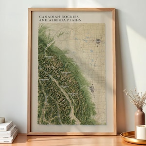 May include: A framed topographic map print titled CANADIAN ROCKIES AND ALBERTA PLAINS, featuring a detailed relief view of mountain ranges in green and brown tones transitioning into the flat, grid-patterned plains of Alberta.