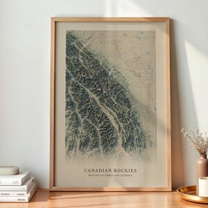 Könnte beinhalten: Ein gerahmter vertikaler Wandkunstdruck mit einer topografischen Karte der kanadischen Rocky Mountains in British Columbia und Alberta. Die Karte zeigt dunkelgrüne Gebirgszüge vor einem hellbeigen Hintergrund mit dem Text CANADIAN ROCKIES.