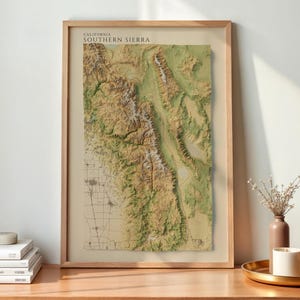 Op de afbeelding: Een ingelijste topografische kaartprint getiteld California Southern Sierra, met een gedetailleerde 3D-reliëfweergave van bergketens en valleien in tinten groen, bruin en wit, gepresenteerd in een lichthouten lijst op een bureau.