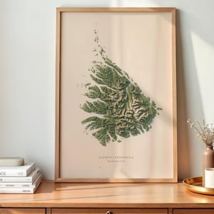 May include: A framed wall art print featuring a detailed, textured green topographical map of the Olympic Peninsula in Washington, set against a light beige background with the text OLYMPIC PENINSULA WASHINGTON printed at the bottom center.