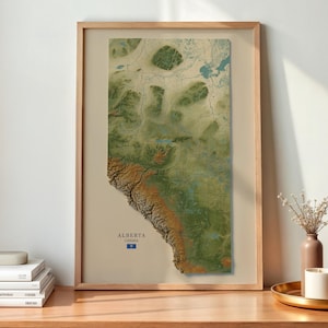May include: A framed vertical map print of Alberta, Canada, featuring a detailed topographical relief design in shades of green and brown. The map is centered within a light wood frame and includes the text ALBERTA CANADA at the bottom.