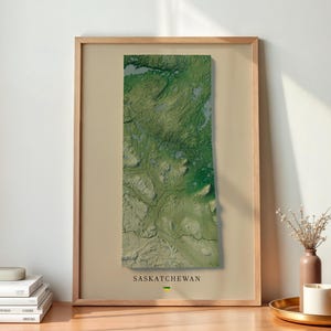 May include: A framed wall art print featuring a satellite-style topographic map of Saskatchewan. The rectangular print is set against a light beige background with the text SASKATCHEWAN printed in black at the bottom center of the frame.