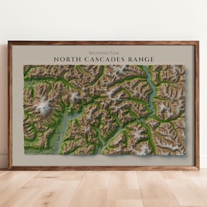 May include: A framed topographic map print of the North Cascades Range in Washington. The map features a detailed 3D-style relief of brown mountain terrain with green valleys and blue water channels, set against a light beige background.