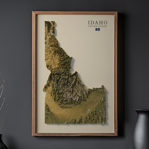 Puede incluir: Un mapa 3D enmarcado de Idaho, EE. UU., con una representación topográfica detallada en tonos verdes y marrones. El mapa está sobre un fondo crema dentro de un marco de madera. El texto "IDAHO UNITED STATES" está en la parte superior.