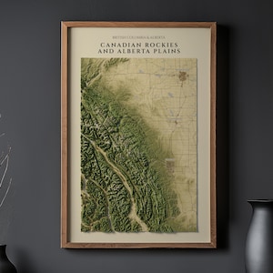 May include: A framed wall map titled British Columbia & Alberta Canadian Rockies and Alberta Plains, featuring a detailed topographical relief of the mountain range and plains in shades of green and tan, enclosed in a simple wooden frame.