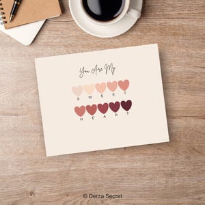 Puede incluir: Una impresi&oacute;n beige con las palabras "You Are My" en escritura sobre una serie de formas de coraz&oacute;n en varios tonos de rosa y rojo. Debajo de los corazones est&aacute;n las letras que deletrean "SWEET HEART". Una taza de caf&eacute; y un bol&iacute;grafo est&aacute;n en el fondo.
