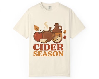 May include: Cream-colored t-shirt featuring a fall-themed graphic. The design includes a red apple, a mug of hot cider with a cinnamon stick, and a jug of cider. The words "CIDER SEASON" are printed in orange and brown.