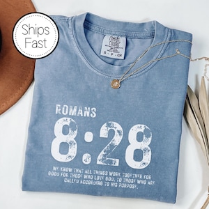 May include: A blue Comfort Colors t-shirt with the text "ROMANS 8:28" in white. The shirt also features a gold necklace with a pendant. The text on the shirt reads, "WE KNOW THAT ALL THINGS WORK TOGETHER FOR GOOD FOR THOSE WHO LOVE GOD, TO THOSE WHO ARE CALLED ACCORDING TO HIS PURPOSE."
