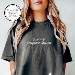 May include: Dark gray t-shirt with the text "Sweat & Scripture Season" in white script. The shirt has a crew neck and short sleeves. A circular graphic in the upper left corner reads "Printed & Shipped in USA".